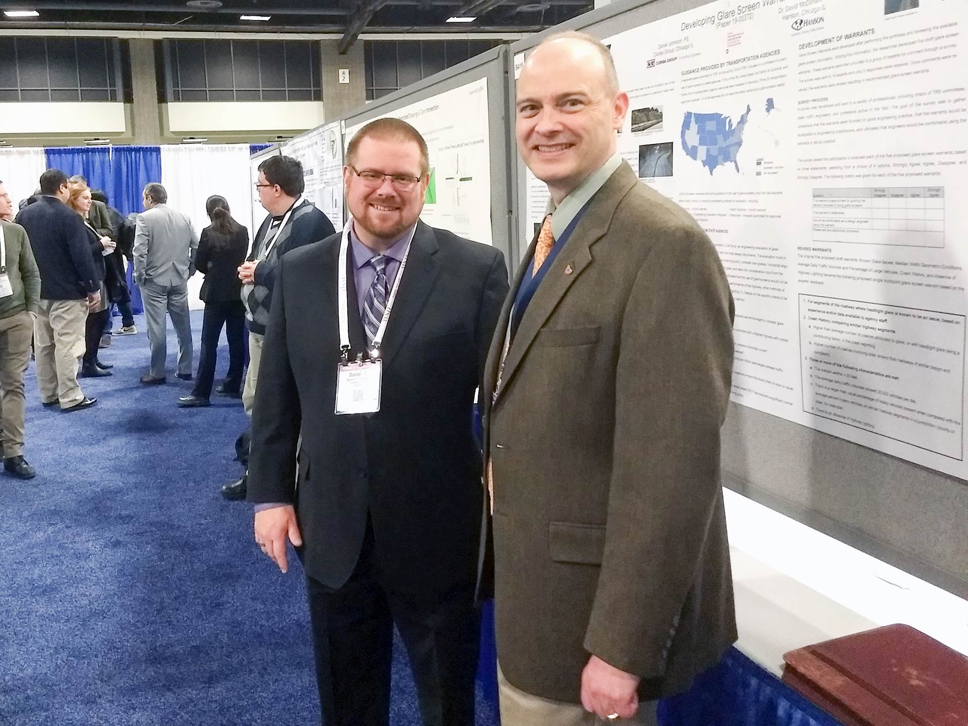 Dan Johnson, in a suit and tie, and David McDonald, in a sport coat and tie, stand together, smiling at the camera, next to a poster of research displayed in a convention hall