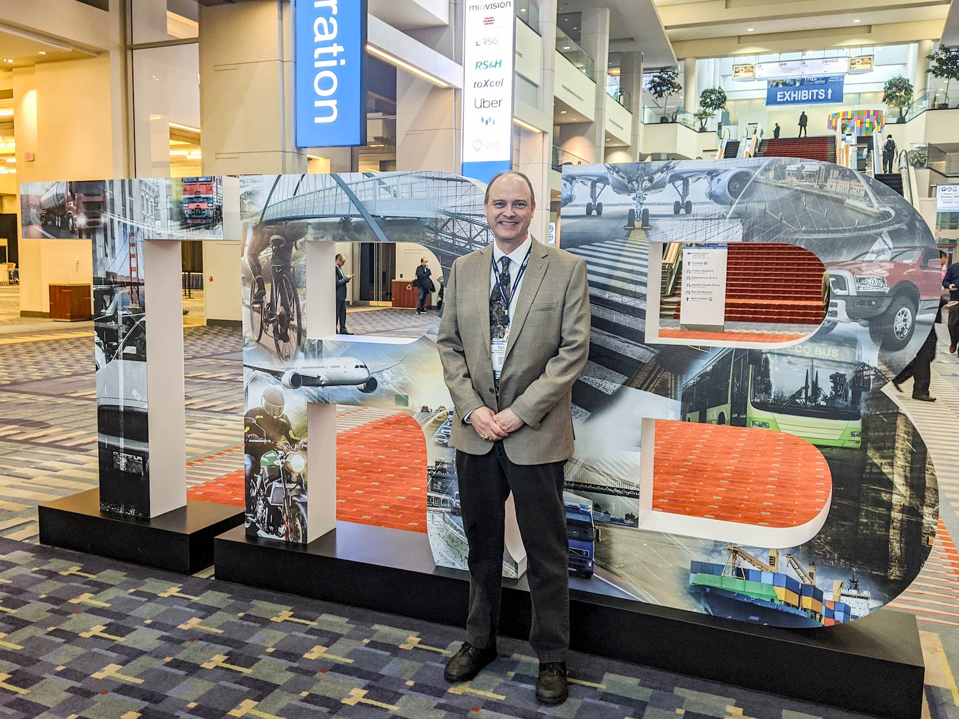 David McDonald, in a sport coat and tie and smiling for the camera, stands in front of large letters spelling TRB at a large event space