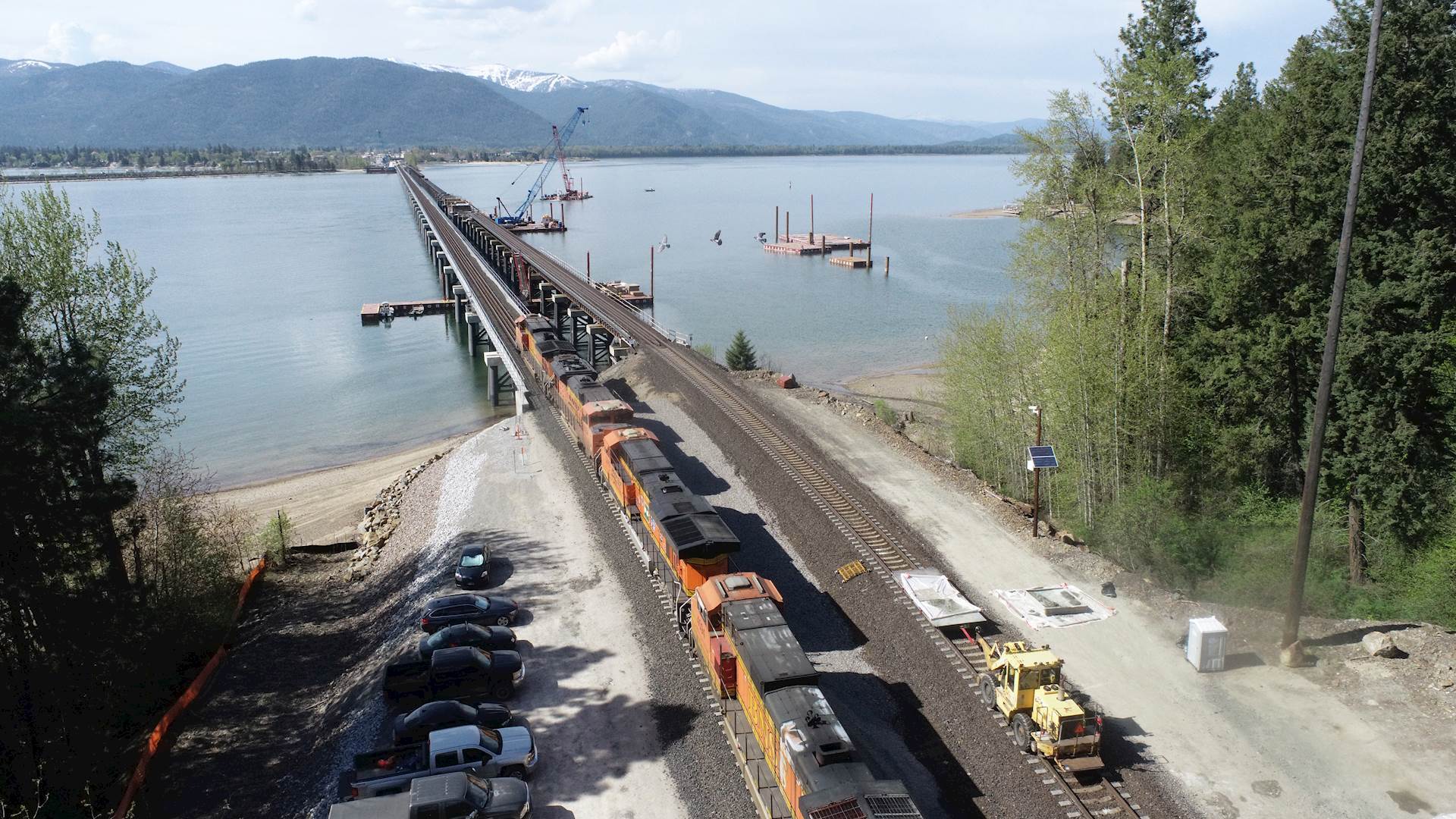 Aerial view of two train tracks leading to their crossing over a lake, with mountains in the background. A train is on one track while the lake bridge of the other track is being constructed. Two construction cranes on barges are on the lake.