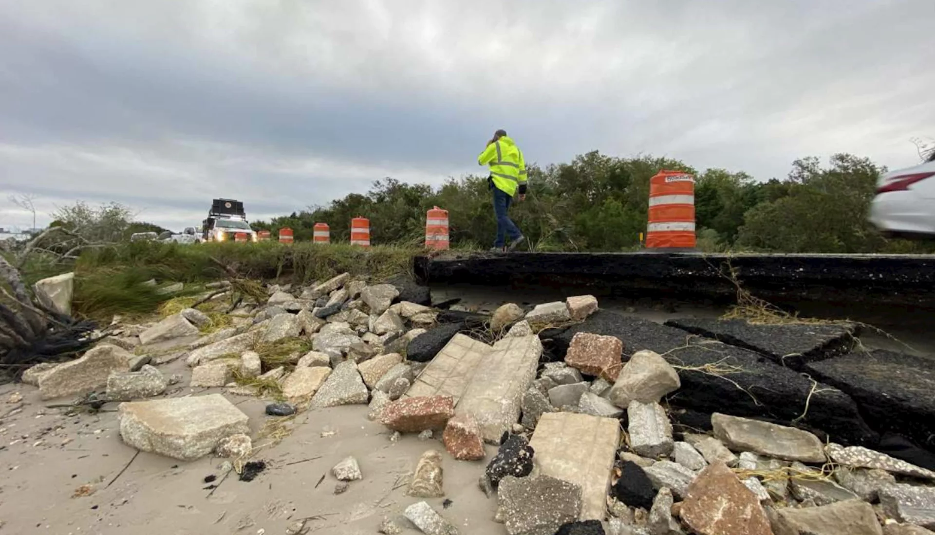 Construction orange cones on the road with a personal in a safety jacket walking; part of the road has broken off and is on the shoreline with other debris and stone; cloudy day.